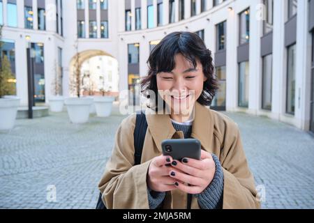 Das stilvolle koreanische Mädchen mit Kopfhörern hört Musik und benutzt Mobiltelefone, steht im Stadtzentrum, wartet auf jemanden auf der Straße und schreibt Textnachrichten Stockfoto