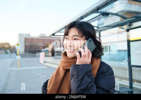 Lächelndes koreanisches Mädchen, das mit dem Handy spricht, an der Bushaltestelle steht, ein Smartphone benutzt, im Winter auf der Straße posiert, in einen Schal eingewickelt, eine schwarze Jacke trägt Stockfoto