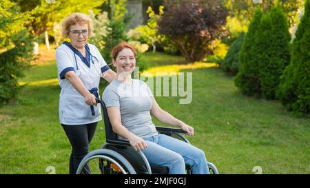 Eine ältere Krankenschwester geht mit einer Frau mittleren Alters im Rollstuhl im Park. Stockfoto