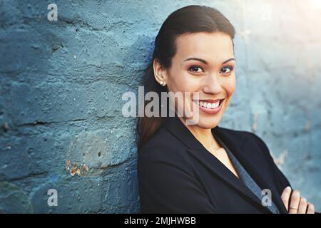 Lassen Sie Ihr Selbstvertrauen durchscheinen. Porträt einer selbstbewussten jungen Geschäftsfrau, die mit gekreuzten Armen gegen eine Mauer posiert. Stockfoto
