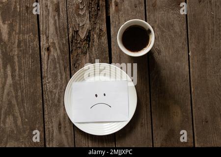 Leerer Teller mit einem traurigen Lächeln auf dem Papier auf einem Holztisch und eine Tasse Kaffee Stockfoto