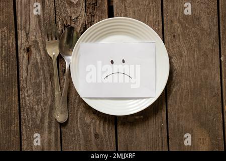 Leerer Teller mit einem traurigen Smiley auf dem Papier und eine Gabel mit einem Löffel auf einem Holztisch, nichts zu essen Stockfoto