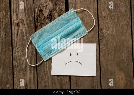 Medizinische Maske trauriges Emoticon auf weißem Papier auf einem Tisch gemalt Stockfoto