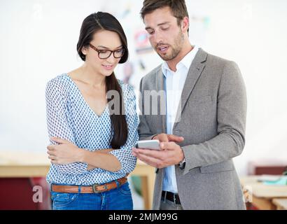Verfolgen Sie Social-Media-Trends. Zwei Kollegen, die sich unterhalten, während sie auf ein Handy schauen. Stockfoto