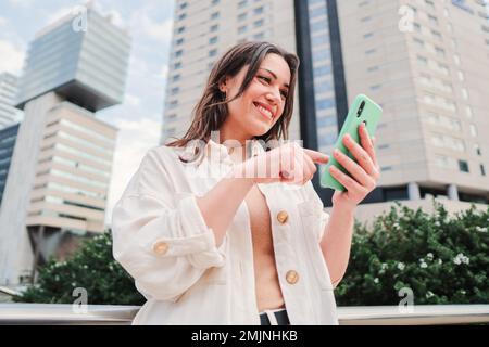 Glückliche junge Frau, die den Bildschirm des Mobiltelefons mit dem Finger berührt, durch die Internet-App scrollt und nach oben wische. Brünette Teenager, die mit dem Smartphone draußen vor Stadtgebäuden lächeln. Hochwertiges Foto Stockfoto