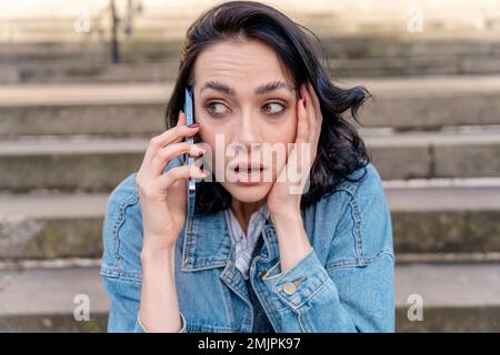 Das Außenporträt einer aufgewühlten jungen Frau in einer Jeansjacke spricht am Telefon über das Lifestyle-Foto Stockfoto
