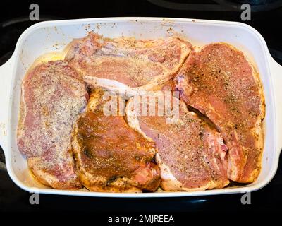 Mehrere rohe Schweinekoteletts mit Gewürzen und Öl in Keramik-Backform auf dem Herd der Küche Stockfoto