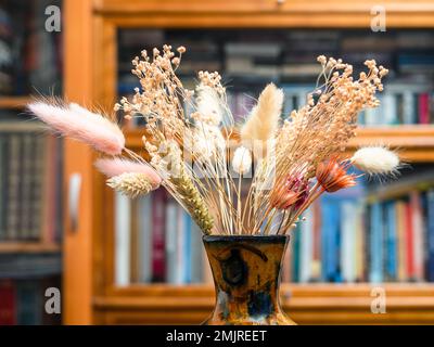 Seitenansicht des Blumenstraußes getrockneter Pflanzen in Vase-Nahaufnahme mit verschwommenem Bücherregal im Hintergrund Stockfoto