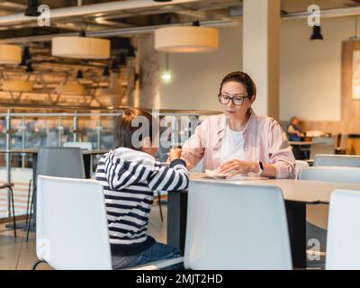 Mutter und Sohn essen im Café. Familie auf dem Food Court im Einkaufszentrum. Frau und Kind essen in einer modernen, nicht wiedererkennbaren Cafeteria zu Mittag. Stockfoto