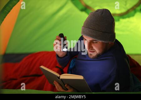 Junger Mann mit Taschenlampe, der im Zelt Buch liest Stockfoto