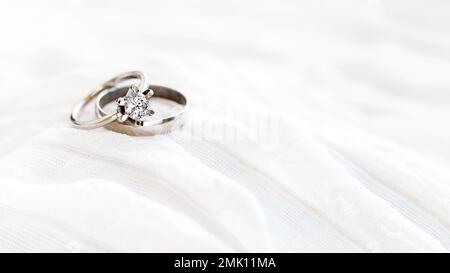 Ein Paar goldene Eheringe auf weißem Textilhintergrund mit Kopierbereich. Verlobungsring mit Diamant auf Textilhintergrund. Symbol für Liebe und Ehe Stockfoto