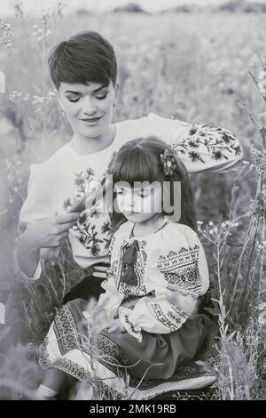 Mom im ukrainischen Nationalkleid flechtet ihre Tochter auf dem Feld Stockfoto