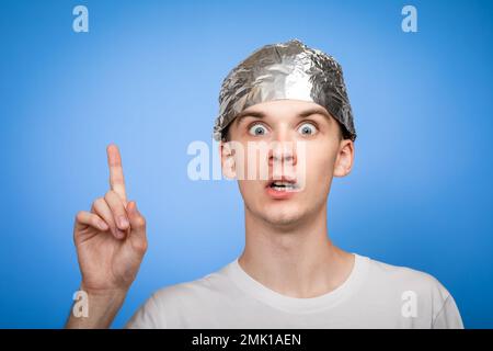 Porträt eines ängstlichen jungen Mannes, der einen Hut aus Alufolie trägt und mit dem Finger nach oben zeigt. Verschwörungstheorien und Paranoya-Konzept. Studio auf blauem Hintergrund aufgenommen Stockfoto