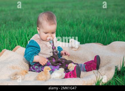 Ein wunderschönes Baby in einem bunten Kleid, das mit Hühnern auf dem Feld sitzt Stockfoto