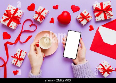 Weibliche Hände halten Smartphone mit Kaffee zum Valentinstag, Geschenkbox und Umschlag, Herzen. Stockfoto