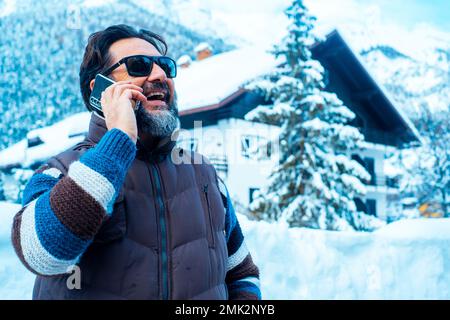 Ein glücklicher, reifer Mann, der am Telefon telefoniert, in einem malerischen Ort im Freien in den Schneebergen, der die Wintersaison und den Urlaub genießt. Positive, schlaue Leute Stockfoto