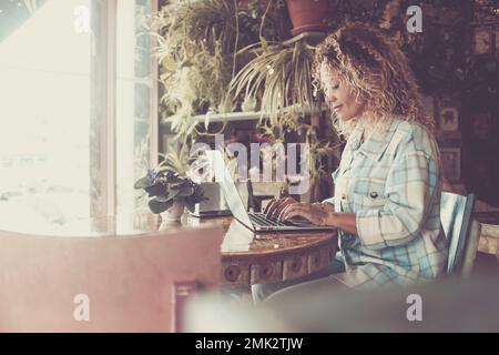 Seitenansicht einer jungen Frau, die am Schreibtisch sitzt und auf dem Laptop schreibt. Eine Geschäftsfrau in moderner, intelligenter Arbeit. Gartenpflanzenladen. Weibliche Menschen Stockfoto
