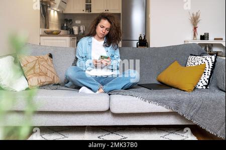 Brünette, lockige Frau in Jeansbekleidung, die zu Hause auf dem Sofa sitzt und ein Handy benutzt. Stockfoto