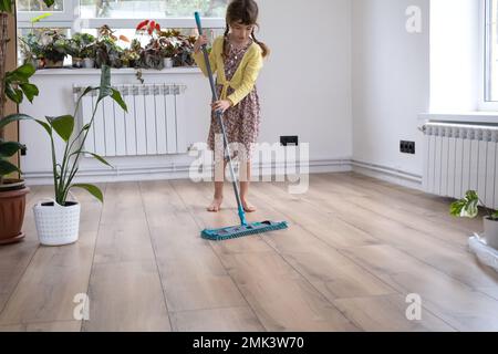 Ein Mädchen tanzt mit einem Mopp, um den Boden in einem neuen Haus zu reinigen - allgemeine Reinigung in einem leeren Raum, die Freude am Umziehen, Hilfe bei der Hausarbeit Stockfoto