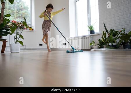 Ein Mädchen tanzt mit einem Mopp, um den Boden in einem neuen Haus zu reinigen - allgemeine Reinigung in einem leeren Raum, die Freude am Umziehen, Hilfe bei der Hausarbeit Stockfoto