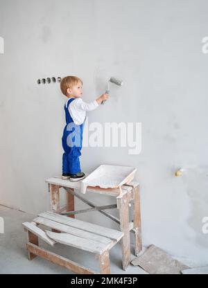 Kleiner Bauarbeiter, der die Wand in einer Wohnung streicht, die gerade renoviert wird. Ein niedliches Kind im Arbeitsanzug, das Malerwalze benutzt, während es zukünftig zu Hause spielt. Stockfoto