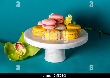 Französische Makkaronkuchen auf einem marmornen weißen Kuchenstand vor blauer Kulisse aus nächster Nähe. Gelbweiße, rosa Makronen. Orchidee und Salbei. Stockfoto