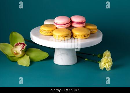 Französische Makkaronkuchen auf einem marmornen weißen Kuchenstand vor blauer Kulisse aus nächster Nähe. Gelbweiß, rosa Makronen. Orchidee und Salbei. Stockfoto