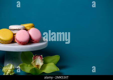 Französische Makkaronkuchen auf einem marmornen weißen Kuchenstand vor blauer Kulisse aus nächster Nähe. Gelbweiße, rosa Makronen. Orchidee und Salbei. Stockfoto