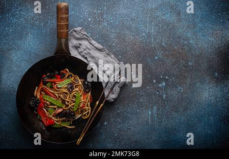 Asiatisches Gericht, gebratene udon-Nudeln mit Gemüse und Pilzen in schwarzer rustikaler Wok-Pfanne mit Holzstäbchen auf rustikalem dunkelblauem Betonhintergrund Stockfoto