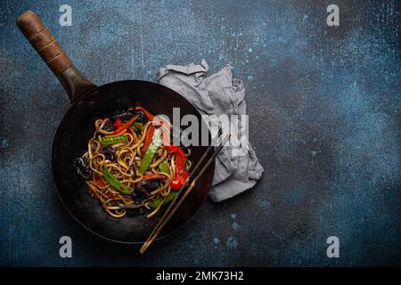 Asiatisches Gericht, gebratene udon-Nudeln mit Gemüse und Pilzen in schwarzer rustikaler Wok-Pfanne mit Holzstäbchen auf rustikalem dunkelblauem Betonhintergrund Stockfoto