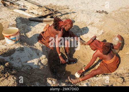 Himba-Frauen sammeln Wasser an einem Wasserloch, am Ufer des trockenen Flussbettes des Hoarusib, Kaokoland, Kunene Region, Namibia Stockfoto