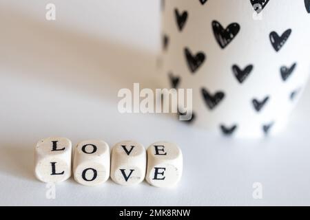 Das Wort "Liebe" besteht aus Holzwürfeln und einem Papierglas mit schwarzen Herzen, isoliert auf weißem Hintergrund Stockfoto