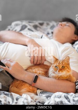 Die süße Ingwerkatze liegt mit ihrem Besitzer im Bett. Der Katzenliebhaber schläft. Die Heilkraft der Haustiere. Kranker Mann mit flauschigem Haustier im Schlafzimmer. Stockfoto