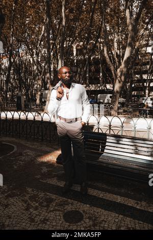 Ein vertikales Bild eines eleganten, gereiften afroamerikanischen Mannes mit Glatze in einem weißen Hemd und einer Brille, mit einem gepflegten schwarzen Bart, der neben einem Pa steht Stockfoto