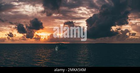 Ein unauffälliger Panoramablick auf einen dramatischen romantischen Sonnenuntergang im Meer mit einer atemberaubenden orangefarbenen Wolkenlandschaft, eine dunkle Silhouette einer luxuriösen Tauchsafari Stockfoto
