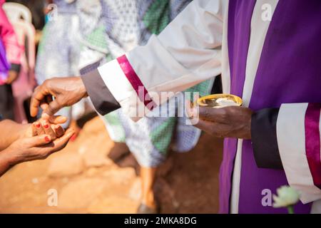 Ein afrikanischer Priester gibt einem Gläubigen sakramentales Brot, konzentriert sich auf die Hand mit sakramentalem Brot Stockfoto