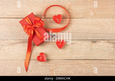 Rote Geschenkbox mit roter Schleife aus Satin und roten Kerzen in Herzform auf Holzhintergrund. Alles gute zum Valentinstag, Muttertag und Geburt Stockfoto