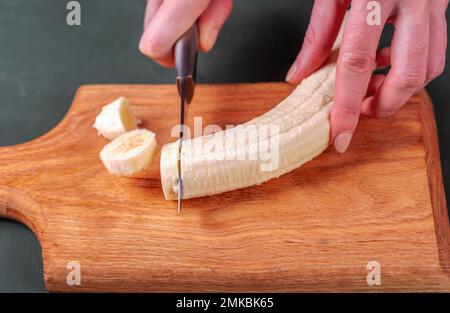 Frau schneidet Banane mit Messer auf Holzbrett in der Küche. Stockfoto
