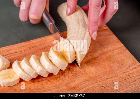 Frau schneidet Banane mit Messer auf Holzbrett in der Küche. Stockfoto