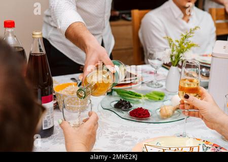 Jüdische Familie feiert das Pessach mit traditionellem Essen am Tisch. Familienessen mit Pessar. Junger Mann gießt Wein in das Glas und Junge liest Haggadah, Frau hebt ein Weinglas Stockfoto