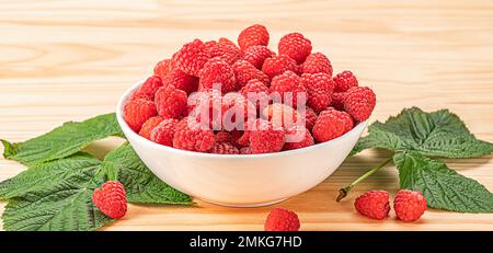 Himbeeren auf weißem Teller auf Holztisch mit Blättern. Bio-Himbeeren ohne den Einsatz von chemischen Düngemitteln und Pestiziden angebaut. Himbeerfrüchte Stockfoto