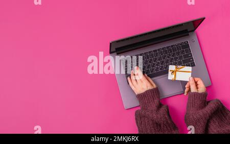 Weihnachtseinkaufskonzept. Flach liegen. Damenhände in einem grünen Pullover auf einer Laptop-Tastatur mit Geschenkkarte auf rotem Hintergrund Stockfoto