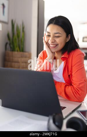 Vertikales Bild einer glücklichen, birassischen Frau, die Videoanrufe auf einem Laptop tätigt und zu Hause lächelt, Kopierbereich. Von zu Hause aus arbeiten, Technologie, Kommunikation und Leben Stockfoto