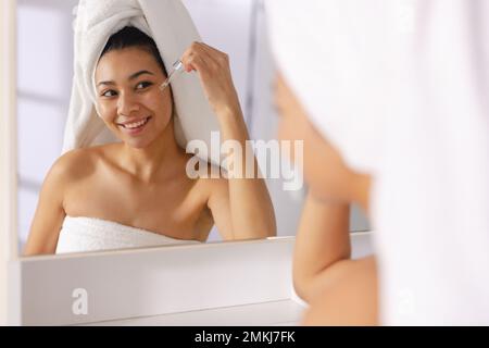 Eine birassische Frau, die ein Handtuch trägt, im Badezimmerspiegel lächelt, ätherische Öle in der Hand hält, Kopierraum. Entspannung, Selbsthilfe, Schönheit, Freizeit und Lebensfreude Stockfoto