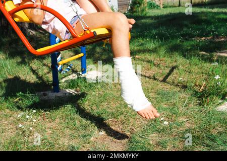 Kinderbeine sind mit Pflaster verbunden - barfuß auf einer Schaukel. Traumatische Ausrüstung, Kinderverletzung, Bluterguss, Verstauchung, Fraktur Stockfoto