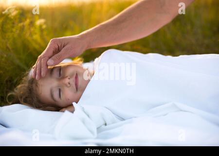 Mädchen schläft auf weißem Bett im Gras, frische Luft. Dads Hand klopft ihm sanft auf den Kopf. Pflege, Schutz, Internationaler Kindertag, Mückenstiche Stockfoto