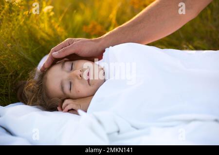 Mädchen schläft auf weißem Bett im Gras, frische Luft. Dads Hand klopft ihm sanft auf den Kopf. Pflege, Schutz, Internationaler Kindertag, Mückenstiche Stockfoto