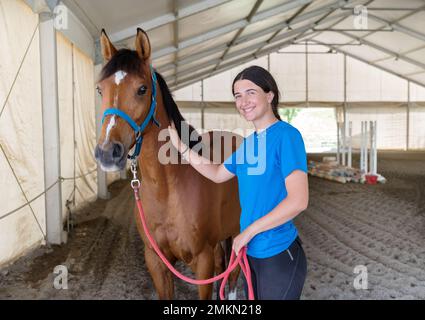 Fröhliche junge Frau mit dunklen Haaren, die mit einem Lächeln in die Kamera schaut und gehorsames Bay Horse nach Training in der Arena am Tag streichelt Stockfoto