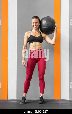 Der ganze Körper einer positiven jungen muskulären Sportlerin mit langen dunklen Haaren in Sportbekleidung, die lächelt und in die Kamera schaut, während sie im Fitnessstudio mit Medicin steht Stockfoto
