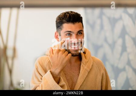 Glücklicher Mann im Bademantel, der lächelt und Creme auf die Wange aufträgt, während er bei der Hautpflege zu Hause in den Spiegel schaut Stockfoto